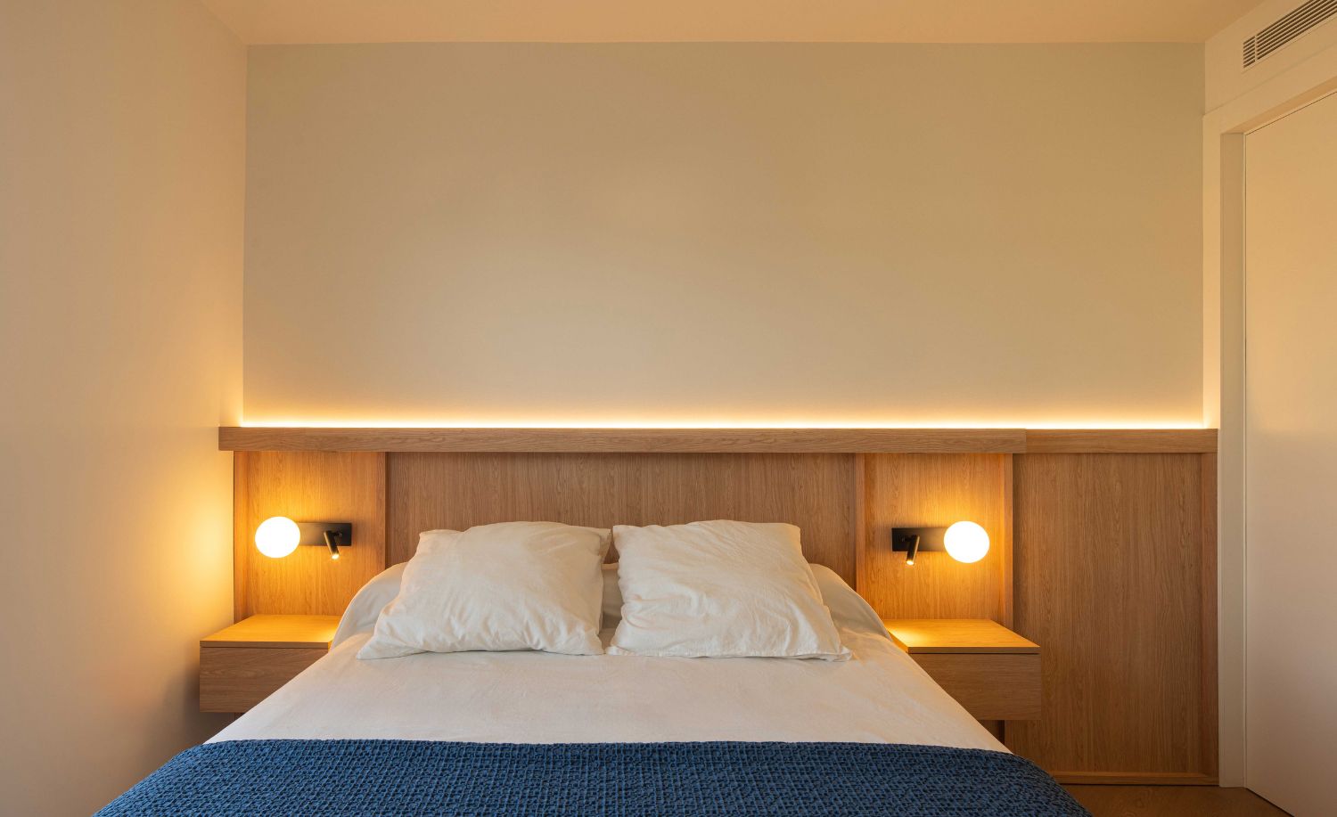 Cama con cabecero de madera retroiluminado y apliques de lectura en el dormitorio reformado