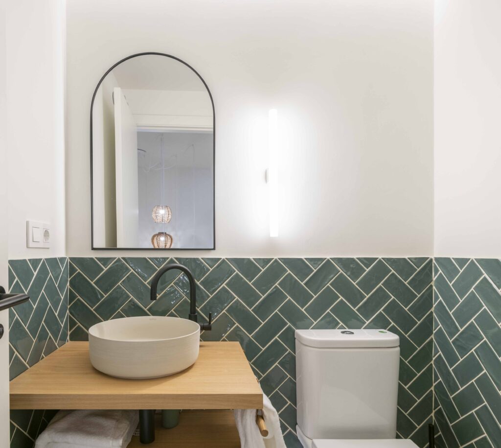 Baño con alicatado verde en espiga, lavabo sobre encimera de madera y espejo arqueado