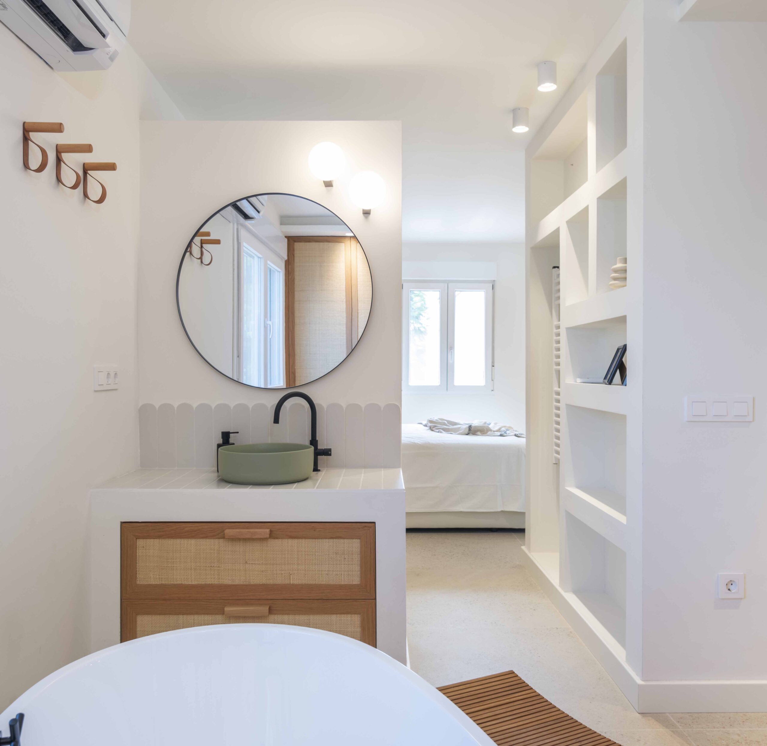 Lavabo con espejo redondo y mueble de ratán junto a la bañera en el baño reformado