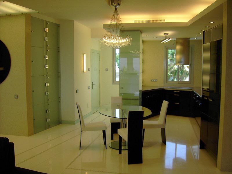 Comedor de cristal con sillas blancas y cocina oscura en la vivienda reformada de Marbella