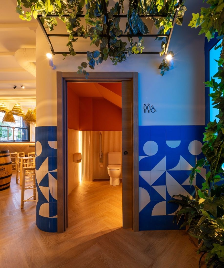 Acceso a los aseos entre plantas colgantes y revestimiento azul geométrico en la cervecería La Oficina
