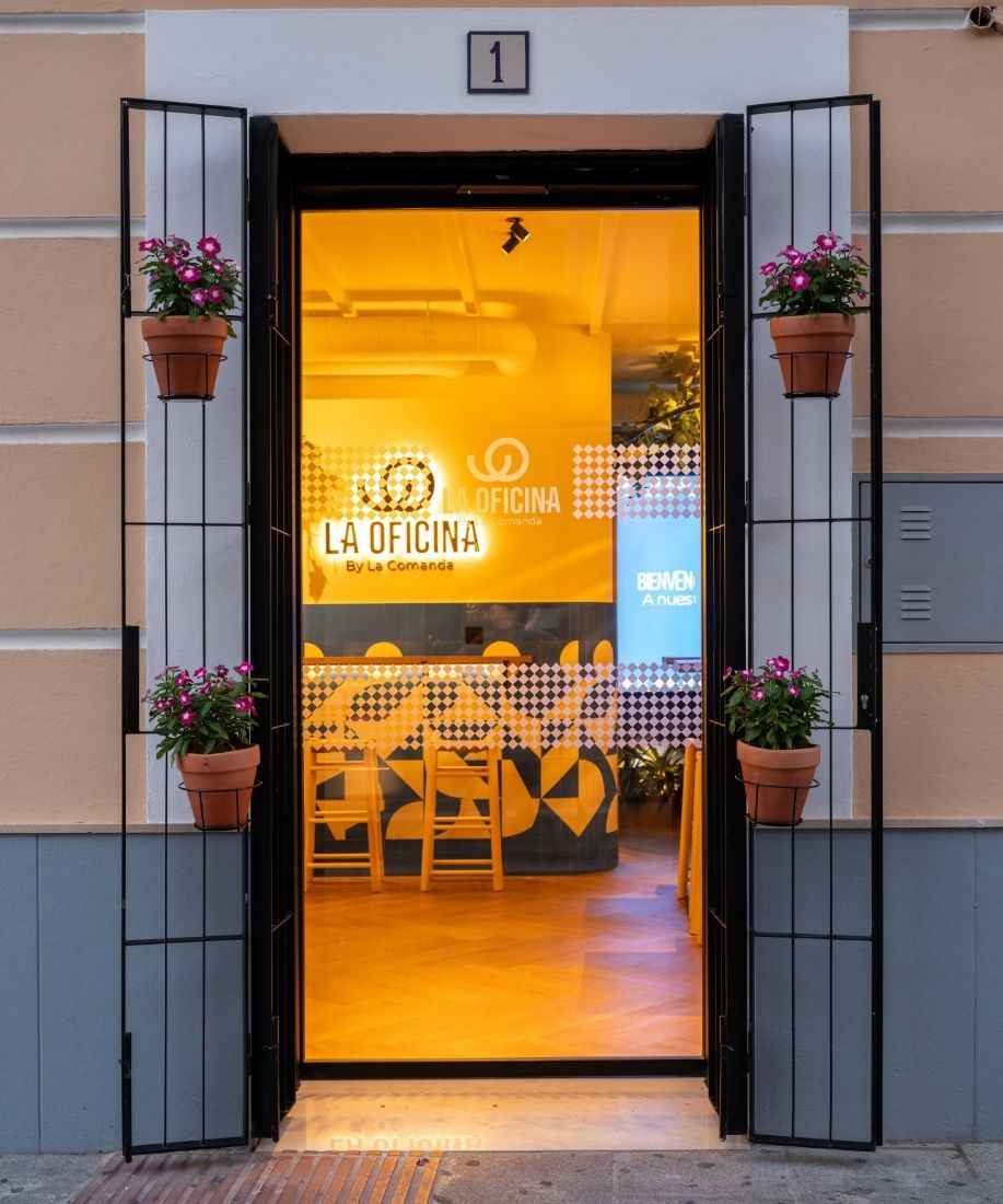 Entrada de la cervecería La Oficina by La Comanda con puertas abiertas y macetas con flores