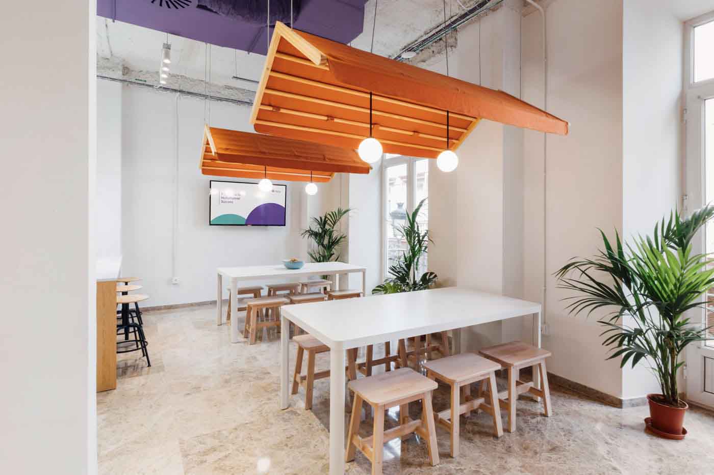 Zona de comedor con mesas largas y plantas bajo paneles naranjas en las oficinas de Plytix