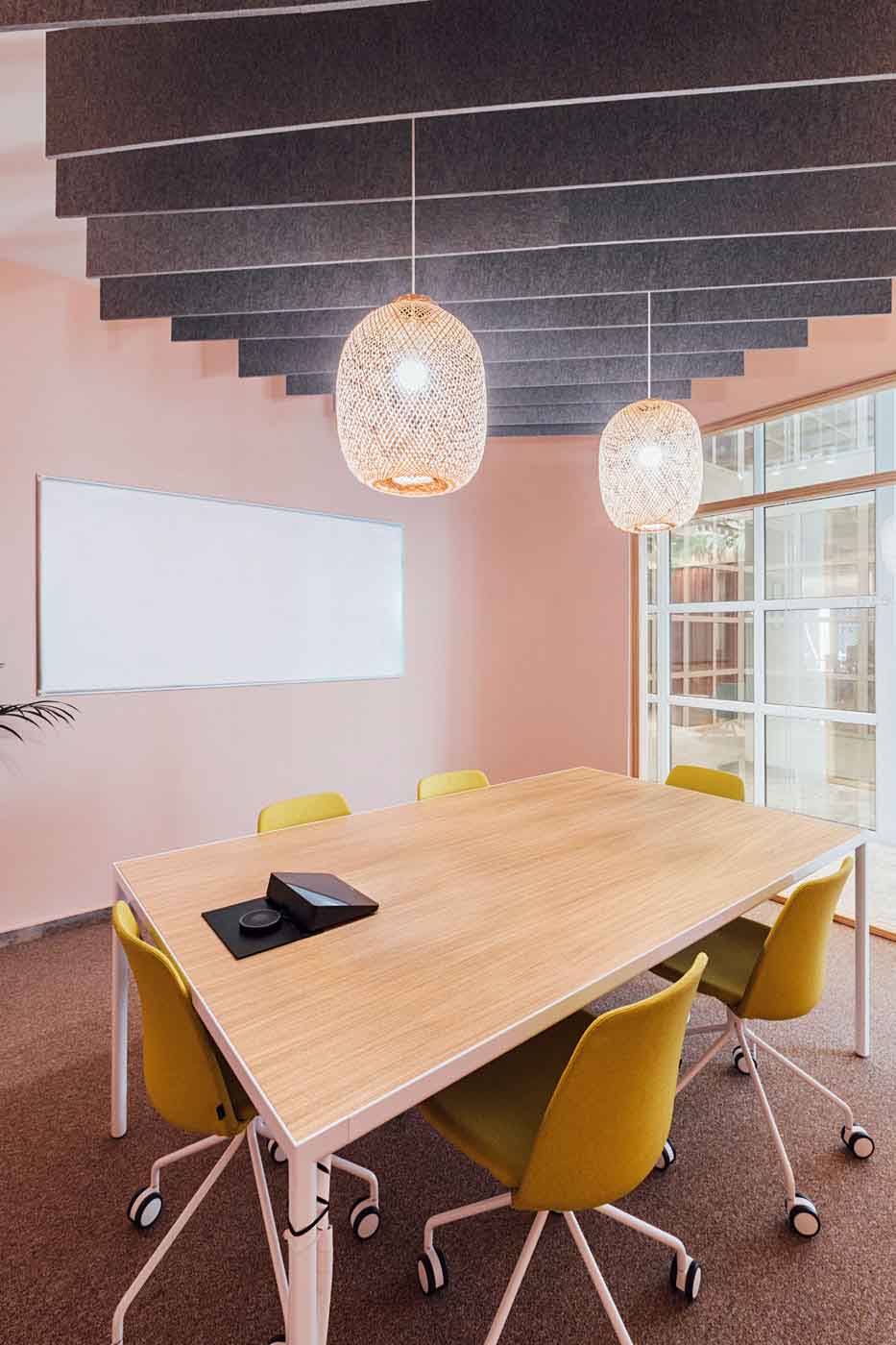 Sala de reuniones de paredes rosadas con mesa de madera, sillas amarillas y lámparas de fibra