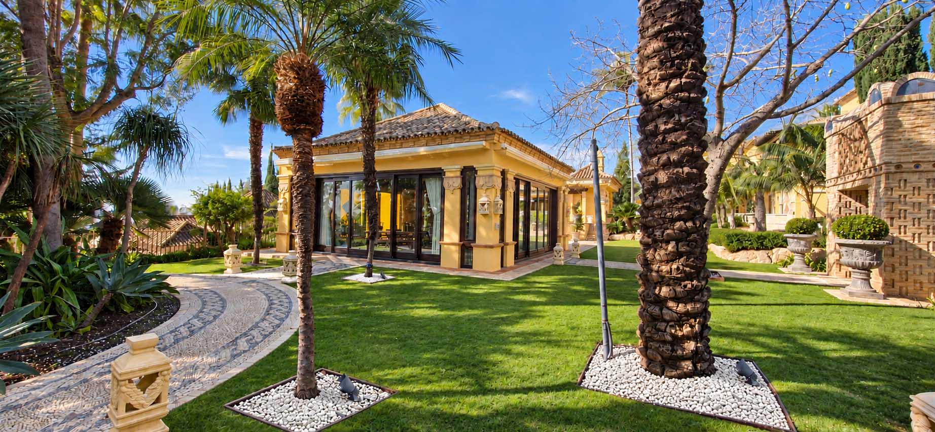 Jardín con césped, palmeras y camino empedrado frente al pabellón de la Villa Casa Blanca